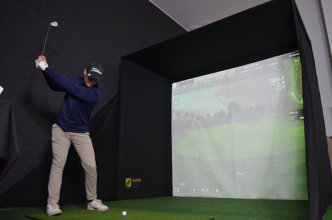 Man swinging a golf club about to hit a ball into the RooBox Golf Simulator Enclosure with projector showing a golf course onto the impact screen.