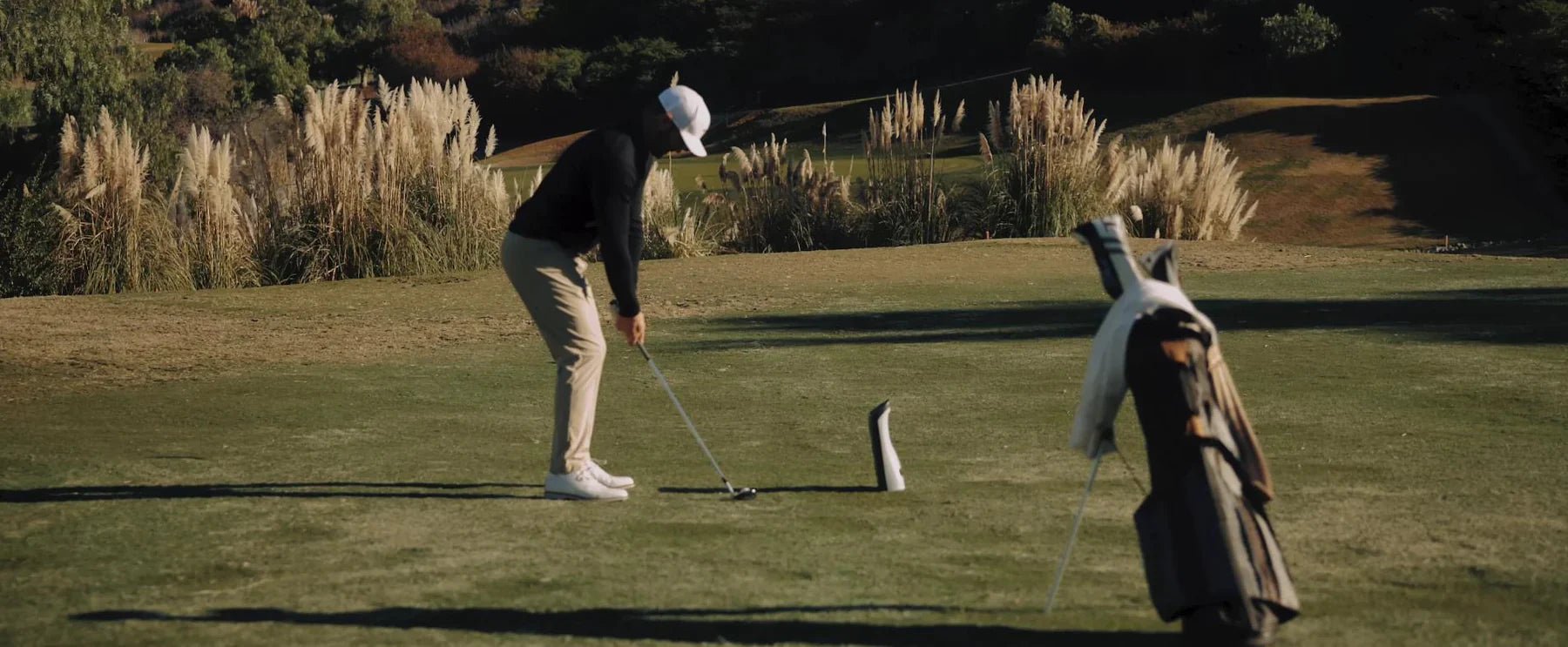Golfer in a white cap and tan pants lining up a putt on a sunny green course with tall golden grasses.