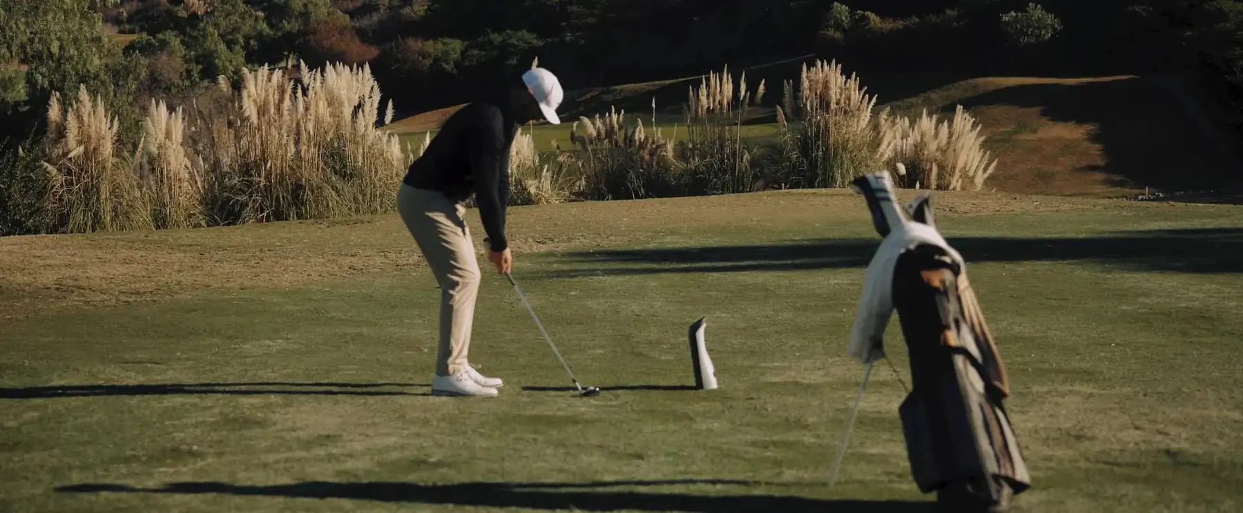 Golfer in a white cap and tan pants lining up a putt on a sunny green course with tall golden grasses.