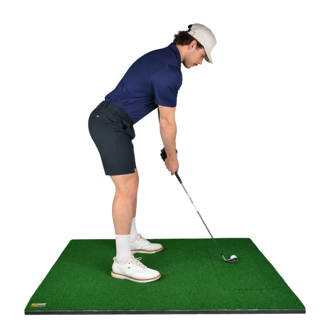 A man in a navy polo shirt, dark shorts, white socks, and a white cap is shown from the back while addressing a golf ball on a green turf hitting mat with a golf club. The background is solid white.