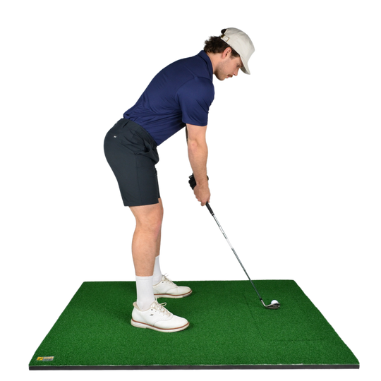 A man in a navy polo shirt, dark shorts, white socks, and a white cap is shown from the back while addressing a golf ball on a green turf hitting mat with a golf club. The background is solid white.