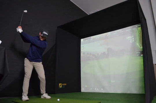 Front-right view of male golfer hitting a ball into `The RooBox` golf simulator enclosure on green turf with golf simulation software projected on the impact screen