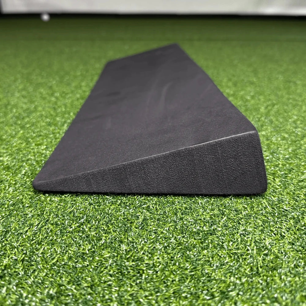 Dark gray EVA foam wedge for golf simulator screen protection, angled on green artificial turf.