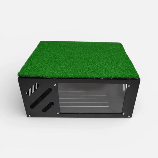 Black metal floor-mounted projector cover with green turf layer on top and clear front shield for golf simulator