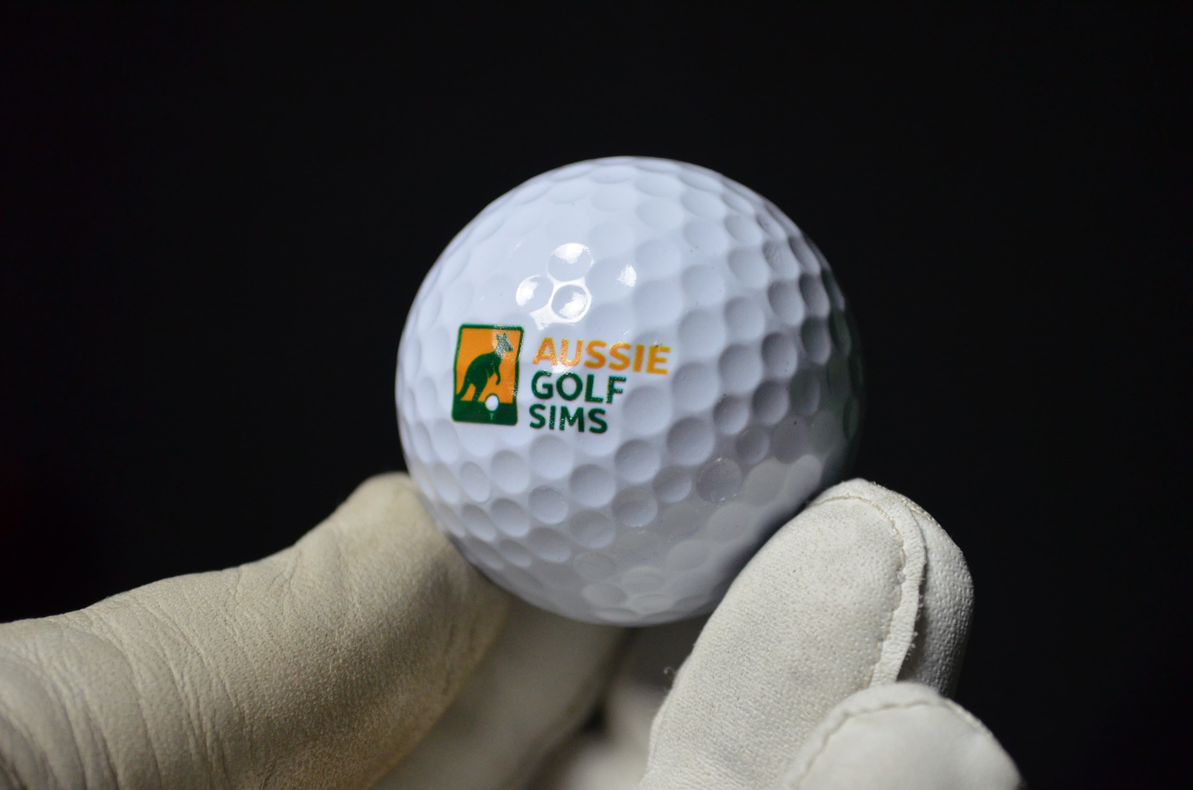 Golf ball with 'Aussie Golf Sims' logo held in a gloved hand against a black background